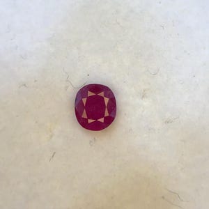 May include: A faceted, oval-shaped ruby gemstone. The gem is a deep, rich red color with internal facets creating a starburst pattern. The ruby is set against a neutral background.
