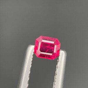 May include: A faceted, rectangular-cut, vibrant red gemstone. The gem is held by a pair of silver tweezers against a dark gray background. The gemstone's facets reflect light, showcasing its clarity and color.