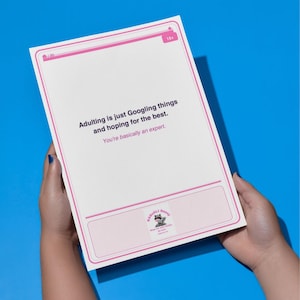 May include: A white card with pink borders and text that reads "Adulting is just Googling things and hoping for the best. You're basically an expert." The card is held against a blue background.
