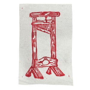 Op de afbeelding: Een rode linosnede van een guillotine op een gebroken witte stof. De guillotine is afgebeeld in een grafische stijl, met een rechthoekige opening en een cirkelvormig gat. De stof heeft een licht getextureerd uiterlijk.