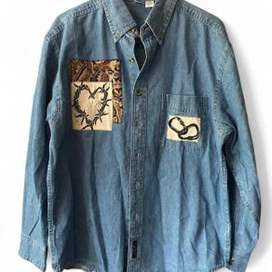 May include: A long-sleeved blue denim button-down shirt featuring two rectangular fabric patches on the chest. The left patch displays a black barbed wire heart design, while the right patch on the pocket shows two interlocking black carabiners.