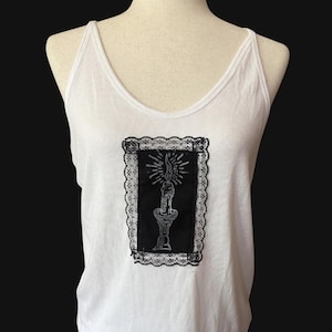 May include: White tank top with a black and white graphic of a candle in a holder, framed by a black lace border. The tank top has a scoop neckline and spaghetti straps.