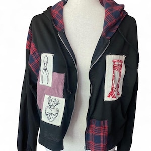 May include: Black zip-up hoodie with red and blue plaid accents on the hood and sleeves. The hoodie features patches with various illustrations, including a pair of scissors, a heart, and a guillotine.