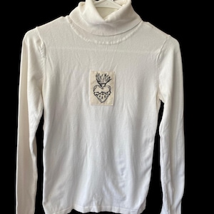 May include: A white turtleneck long-sleeve shirt. A rectangular patch with a black embroidered heart design is centered on the chest. The heart has a crown of flames and a thorny border. The shirt is made of a soft, knit fabric.
