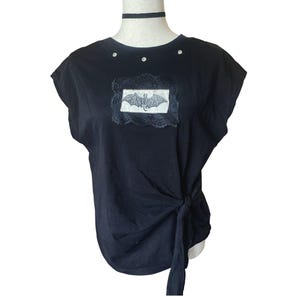 Può includere: T-shirt nero con grafica a pipistrello. La maglietta presenta una toppa decorativa in pizzo con un disegno di pipistrello ed è impreziosita da strass. La maglietta è annodata sul lato e include un choker nero.