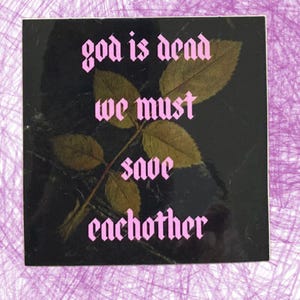 May include: Square sticker with the text "god is dead we must save eachother" in pink gothic font. The sticker has a black background with a leaf and stem design in the center.