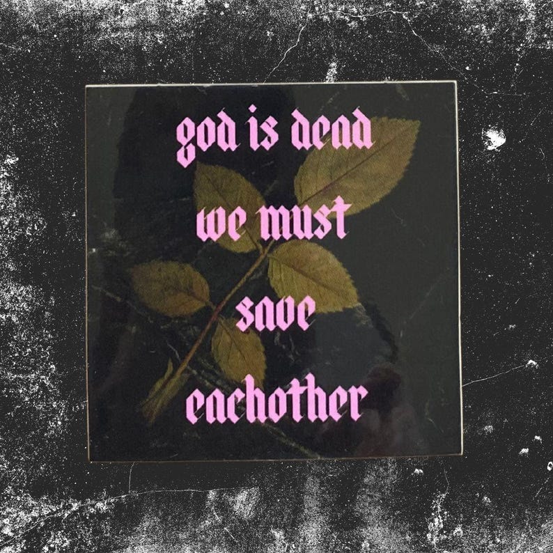 May include: Square black tile with the text "god is dead we must save each other" in pink gothic font. A sprig of leaves with brown foliage is centered on the tile.