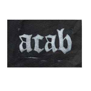 以下が含まれることがあります： 黒い布のパッチに、白いスタイリッシュな文字で「ACAB」と書かれている。