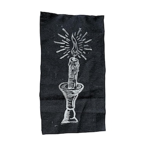 May include: A white candle with a flame on top, sitting on a black candlestick with a decorative base. The candle is on a black fabric background.