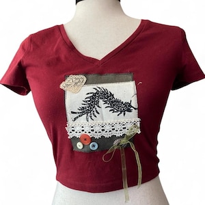 May include: A maroon V-neck t-shirt featuring a rectangular fabric patch on the chest. The patch displays a black centipede illustration, white lace trim, a small beige lace piece, three colorful buttons, and a thin olive green ribbon bow.