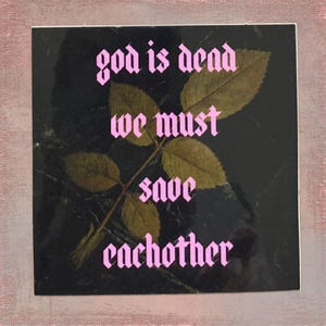 May include: Square sticker with the text "god is dead we must save each other" in pink gothic font. A sprig of dried leaves and a stem are in the center, set against a dark background.