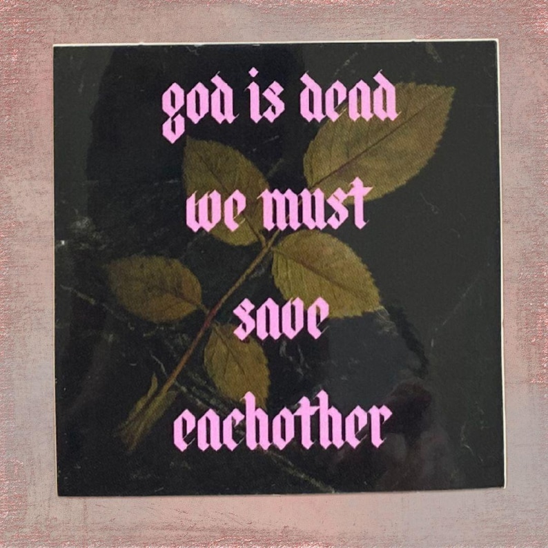 May include: Square sticker with the text "god is dead we must save each other" in pink gothic font. A sprig of dried leaves and a stem are in the center, set against a dark background.