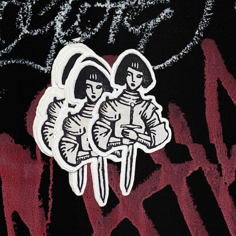 May include: A stack of white stickers featuring a black and white illustration of a person in armor holding a sword. The stickers are on a black background with red and white graffiti.