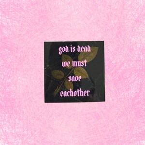 May include: Square black art print with the text "god is dead we must save eachother" in pink gothic font. The print features a sprig of brown leaves. The background is a textured pink.