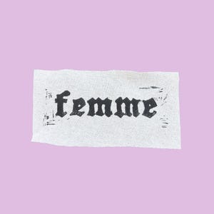 Op de afbeelding: Een rechthoekig stuk getextureerd, gebroken wit papier met het woord "femme" in zwarte, gotische letters. Het papier staat tegen een zachte, lavendelkleurige achtergrond.