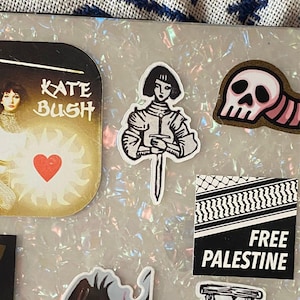 May include: A collection of stickers featuring various designs. One sticker has the text "KATE BUSH" with a heart. Another depicts a figure holding a sword. A third sticker shows a skull. A fourth sticker has the text "FREE PALESTINE".