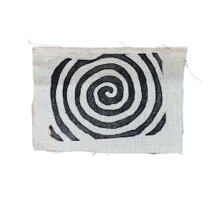 Puede incluir: Un parche de tela blanco con un diseño de espiral negro. La espiral está centrada en el parche y está formada por círculos concéntricos.