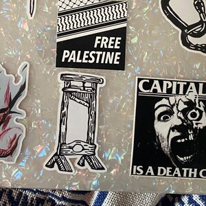 May include: A collection of black and white stickers on a holographic background. The stickers feature various designs, including a guillotine, the text "FREE PALESTINE," and a screaming face with the words "CAPITAL IS A DEATH CULT."
