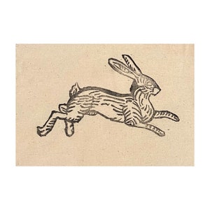 May include: A black and white illustration of a running rabbit on a beige background. The rabbit is shown in profile, with its ears up and legs extended in mid-leap. The artwork has a rustic, hand-drawn aesthetic, reminiscent of a woodcut.