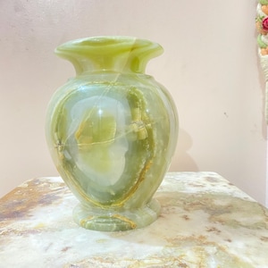 Könnte beinhalten: Eine grüne Onyx-Vase mit ausgestelltem Oberteil und rundem Körper. Die Vase hat eine polierte Oberfläche und ein natürliches, marmoriertes Muster mit Grün- und Goldtönen. Sie steht auf einer Steinoberfläche.