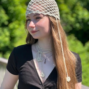 May include: A cream-colored crochet head covering featuring an open-work net pattern with a scalloped edge. The accessory includes long, thin crocheted ties that hang down, each finished with a small, delicate crocheted flower at the end.