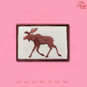 May include: Embroidered patch featuring a brown moose silhouette on a white background, framed by a dark brown border. The patch is rectangular and suitable for application on clothing or accessories.