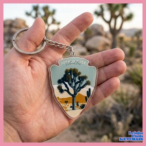 May include: A keychain featuring a desert landscape with a Joshua Tree illustration. The keychain has a silver metal ring and chain. The text "Joshua Tree National Park" is printed at the top. The keychain is held in a person's hand.