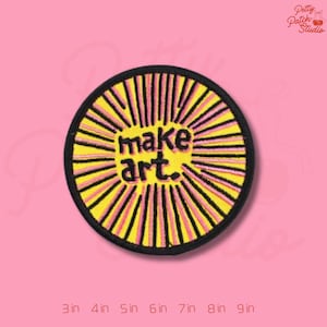 May include: Embroidered patch with the text "make art." The patch is circular, featuring a yellow center with radiating pink and yellow lines, and a black border. The background is pink.
