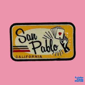 May include: A rectangular patch with a black border and a yellow background. The patch features the words "San Pablo" in black script, with "CALIFORNIA" below. A hand holds playing cards with a heart.