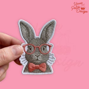 May include: Embroidered patch featuring a gray rabbit wearing red glasses and a bow tie. The patch has a white border and is held against a pink background. The text "Uzura Patch Design" is in the upper right corner.