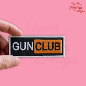 May include: A rectangular patch with the words "GUN CLUB" in white capital letters on a black background. The word "CLUB" is on an orange square. The patch is held against a pink background.