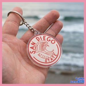 May include: A clear, round keychain with a silver chain and ring. The keychain features the words "San Diego State Aztecs" in red, with a wave graphic. The keychain is held in a hand.