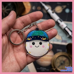May include: A clear acrylic keychain featuring a cartoon character with a smiling face, wearing a blue and green hat with a starry band. The keychain has a silver chain and ring. The background includes a map and a model rocket.