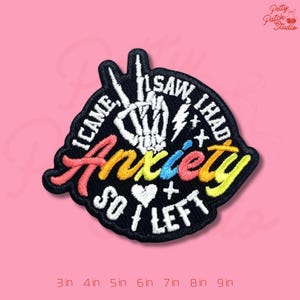 May include: Embroidered patch with the text "I came, I saw, I had anxiety so I left." The word "Anxiety" is in rainbow colors. A white skeleton hand is in the center. The patch is black with a pink background.
