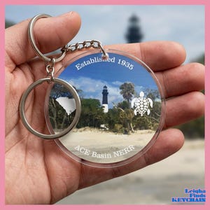 May include: A clear acrylic keychain with a silver keyring. The keychain features a scenic image of a lighthouse, beach, and a turtle, with the text "Established 1935" and "ACE Basin NERR". The South Carolina outline is also visible.