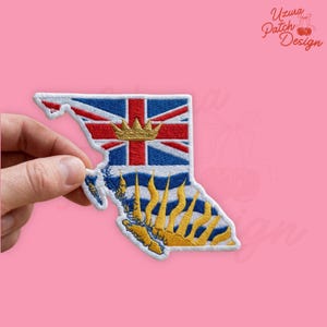 May include: Embroidered patch in the shape of British Columbia, Canada, featuring the provincial flag and a gold crown. The patch has a white border and is held against a pink background. The text "Uzura Patch Design" is in the upper right corner.