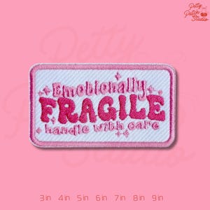 May include: A rectangular embroidered patch with a white background and pink border. The patch features the words "Emotionally FRAGILE handle with care" in pink lettering. The patch is set against a pink background.