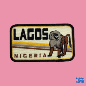 May include: Embroidered patch featuring the word "LAGOS" in black block letters above "NIGERIA". A baboon illustration is on the right. The patch has a black border with yellow and brown accents, set against a cream background.