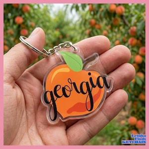 May include: A clear acrylic keychain in the shape of a peach, with the word "georgia" in black script. The peach is orange with a green leaf. The keychain is held in a hand against a blurred background of peach trees.
