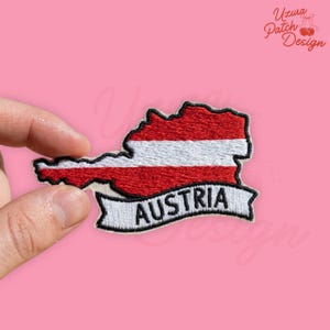 May include: Embroidered patch in the shape of Austria, featuring the country's flag colors: red, white, and red. The word "AUSTRIA" is embroidered on a white banner below the map. The patch is held against a pink background.