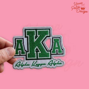 May include: Embroidered patch featuring the letters "AKA" in green with a white border. The words "Alpha Kappa Alpha" are embroidered in green script below the letters. The patch is held against a pink background.