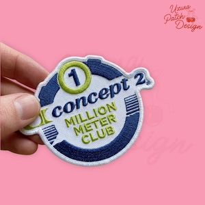 May include: Embroidered patch with the text "Concept 2 Million Meter Club". The patch is white with blue and green accents. The number "1" is in a green circle. The patch is held against a pink background.