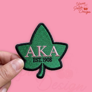 May include: A green embroidered patch in the shape of a leaf with the letters "AKA" in pink and the text "EST. 1908" in green. The patch has a black border and is held against a pink background.