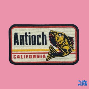 May include: Embroidered patch featuring a fish illustration with the text "Antioch California". The patch has a rectangular shape with a black border, a cream-colored background, and yellow and red stripes.