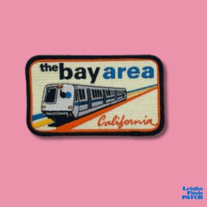 May include: Embroidered patch featuring a train and the words "the bay area" in white and blue, with "California" in orange. The patch has a black border and is set against a pink background.