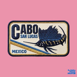 May include: Embroidered patch featuring a sailfish design with the text "CABO SAN LUCAS MEXICO." The patch has a cream background with navy blue and gold accents. The sailfish is navy blue with gold detailing.