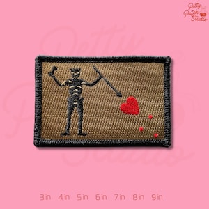May include: Embroidered patch featuring a black skeleton figure holding a spear and a flag with a red heart. The patch has a tan background with a black border. The patch measures 3 inches by 2 inches.