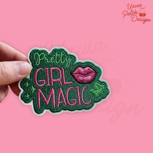 May include: Embroidered patch with the words "Pretty Girl Magic" in pink and green. The patch features a pair of pink lips and green accents. The patch is held against a pink background.