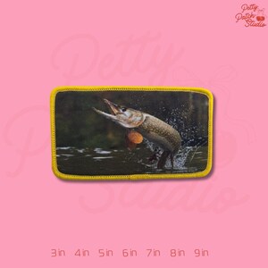 May include: Embroidered patch featuring a leaping pike fish with its mouth open, set against a water and forest backdrop. The patch has a yellow border. The text "Petty Patch Studio" is in the upper right corner.