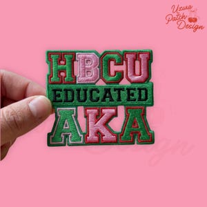 May include: Embroidered patch with the words "HBCU EDUCATED AKA" in green, pink, and black. The patch has a layered design with a pink background. The patch is held by a hand.